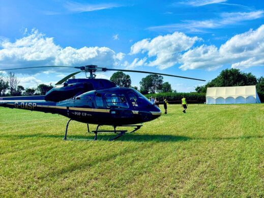 Black helicopter parked on a grassy field with people in the background and a white marquee under a blue sky.