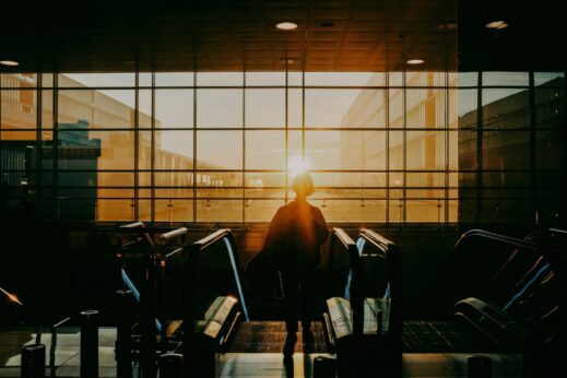 Person stands on an escalator in a darkened terminal, facing a bright sunset through large glass walls.